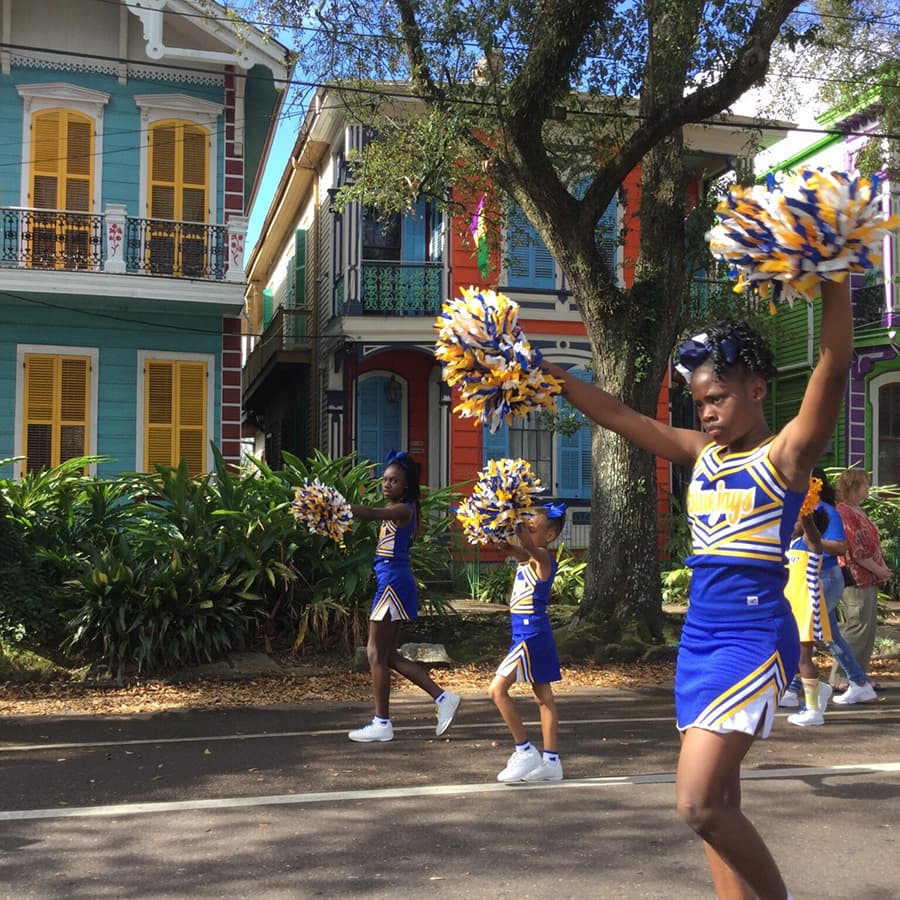Cheerleaders in blue and gold uniforms perform with pom-poms in front of colorful houses during a parade.