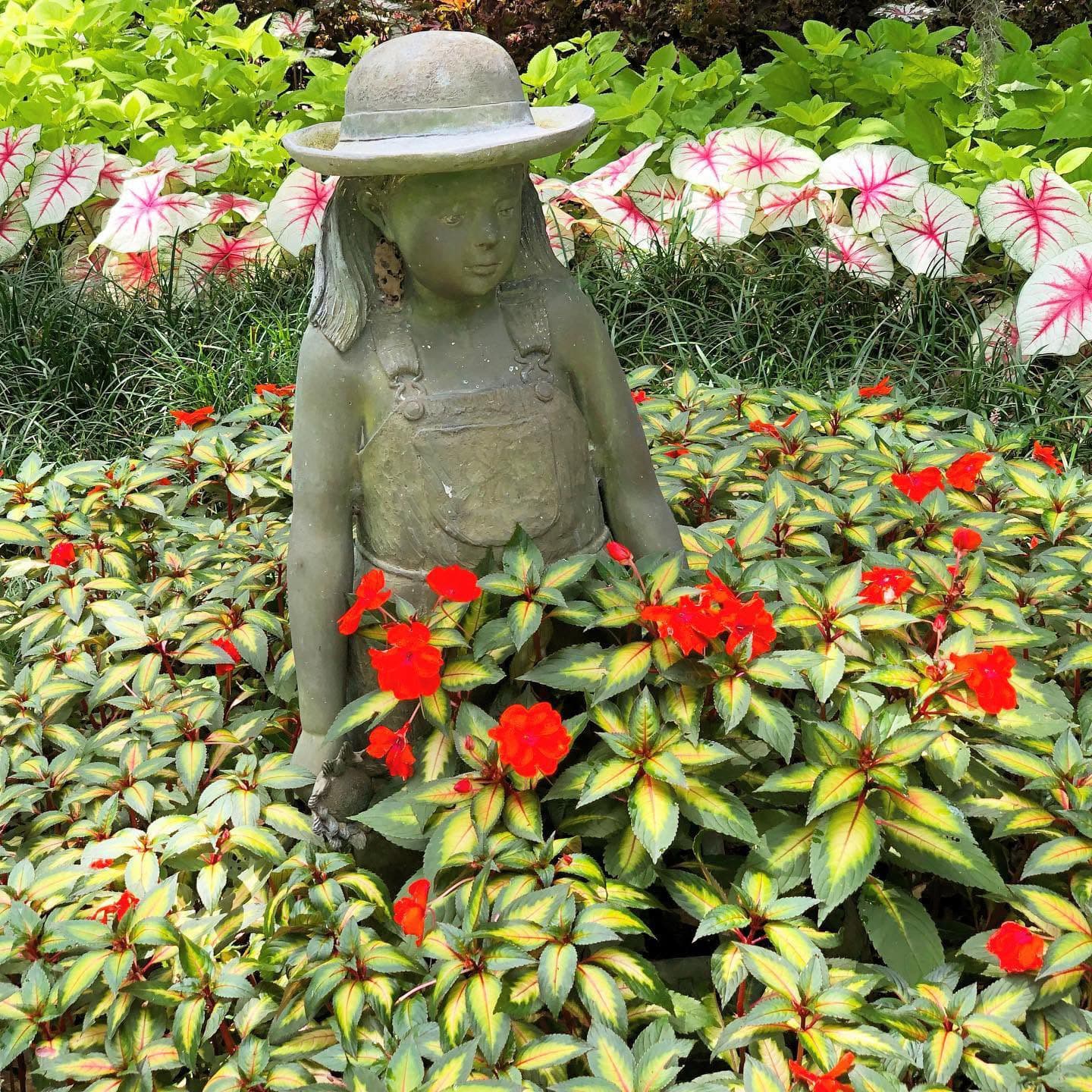 A stone statue of a girl wearing a hat and overalls is surrounded by vibrant red flowers and green foliage.