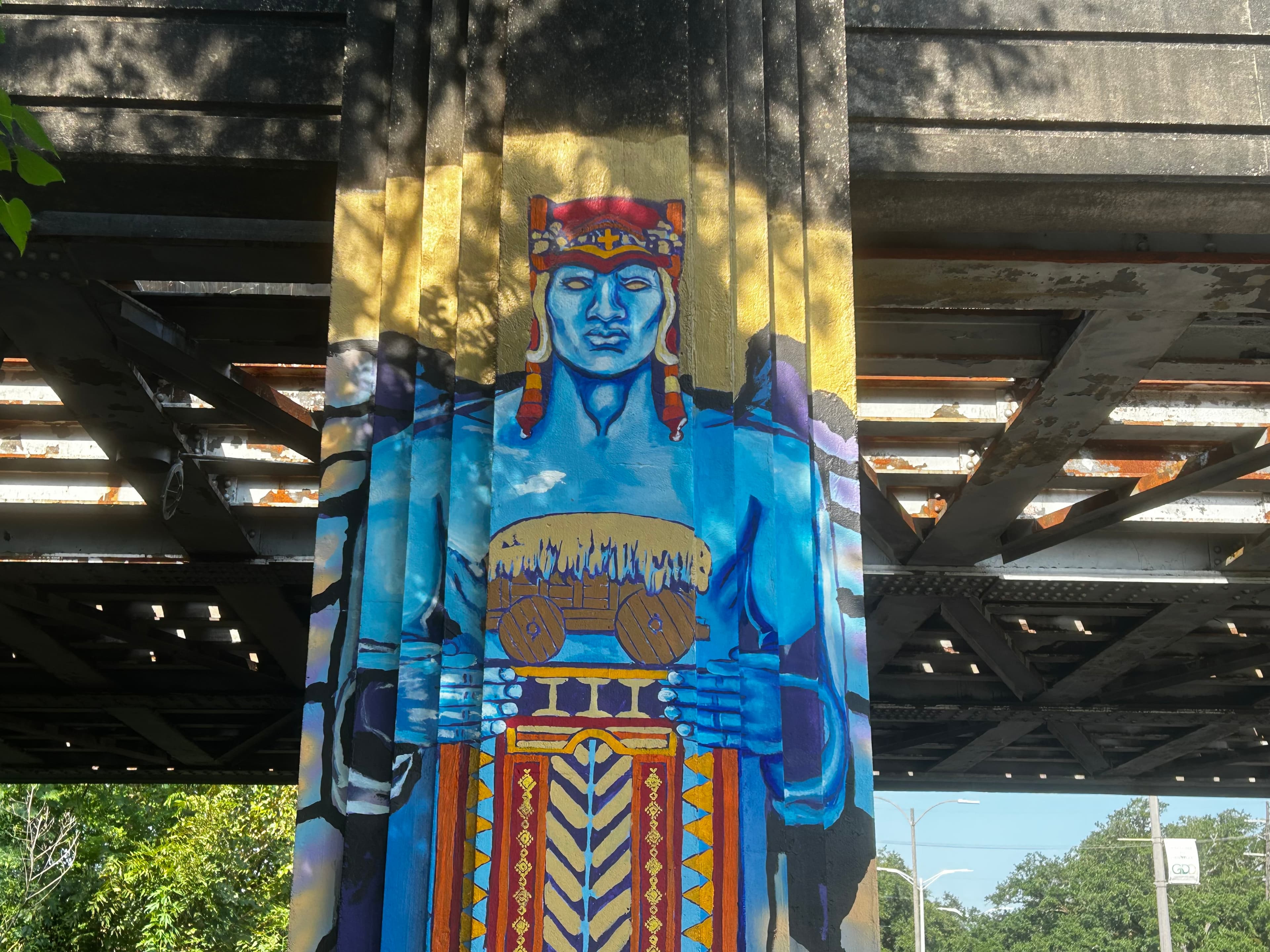 Colorful mural of a blue figure holding a drum, located on a pillar under a bridge.
