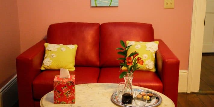 A red couch with decorative yellow pillows sits beside a marble table adorned with a vase, a tissue box, and a plate of snacks.