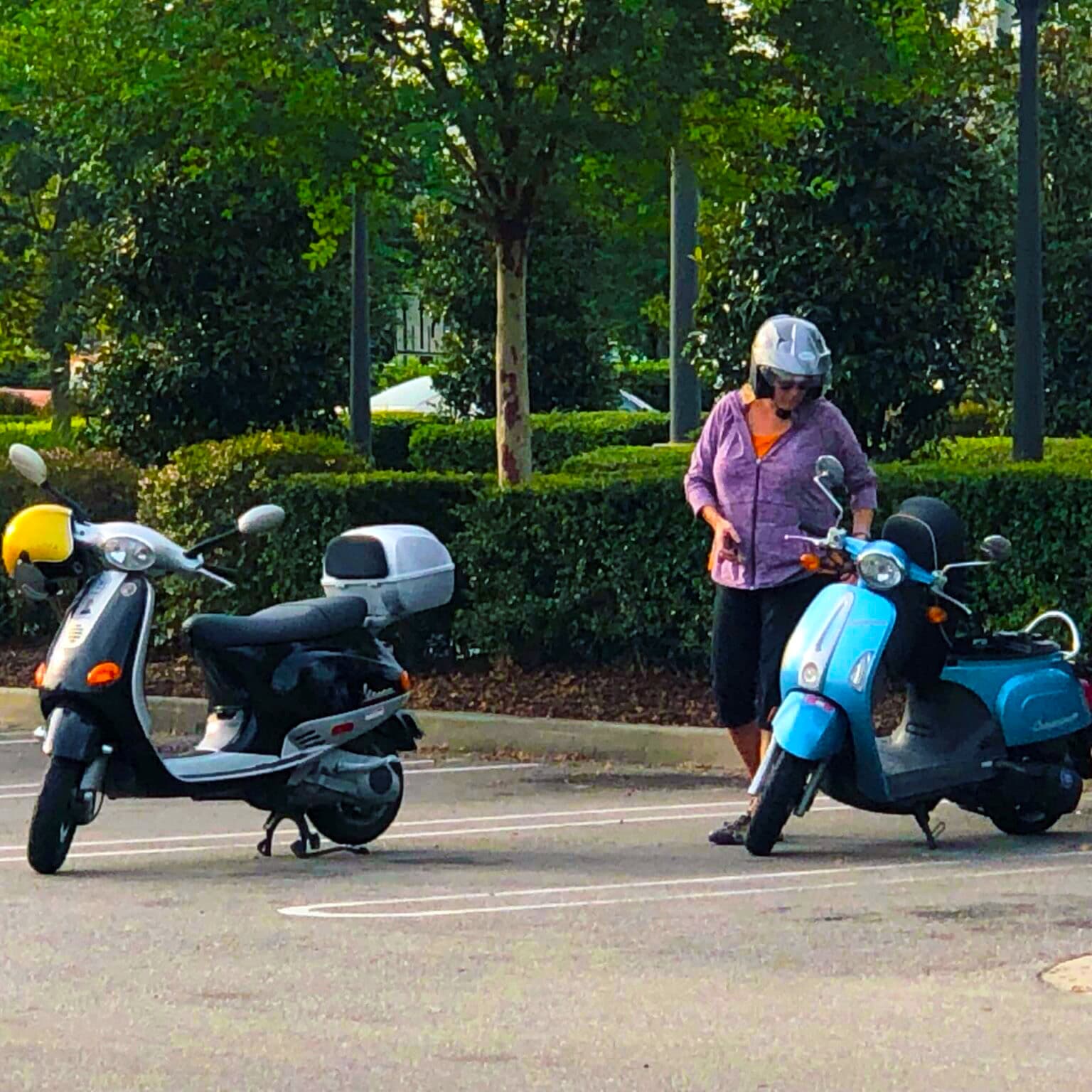 A woman in a helmet stands between two parked scooters in a lush, green setting.