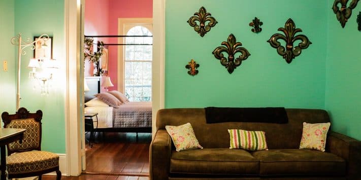 A cozy living area featuring a turquoise wall, a brown couch with patterned cushions, and a glimpse of a pink bedroom through an open doorway.