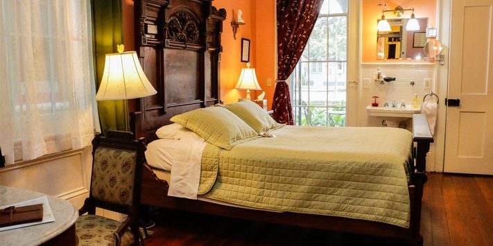 A cozy bedroom featuring a wooden bed, soft green bedding, ambient lighting, and a window with curtains.