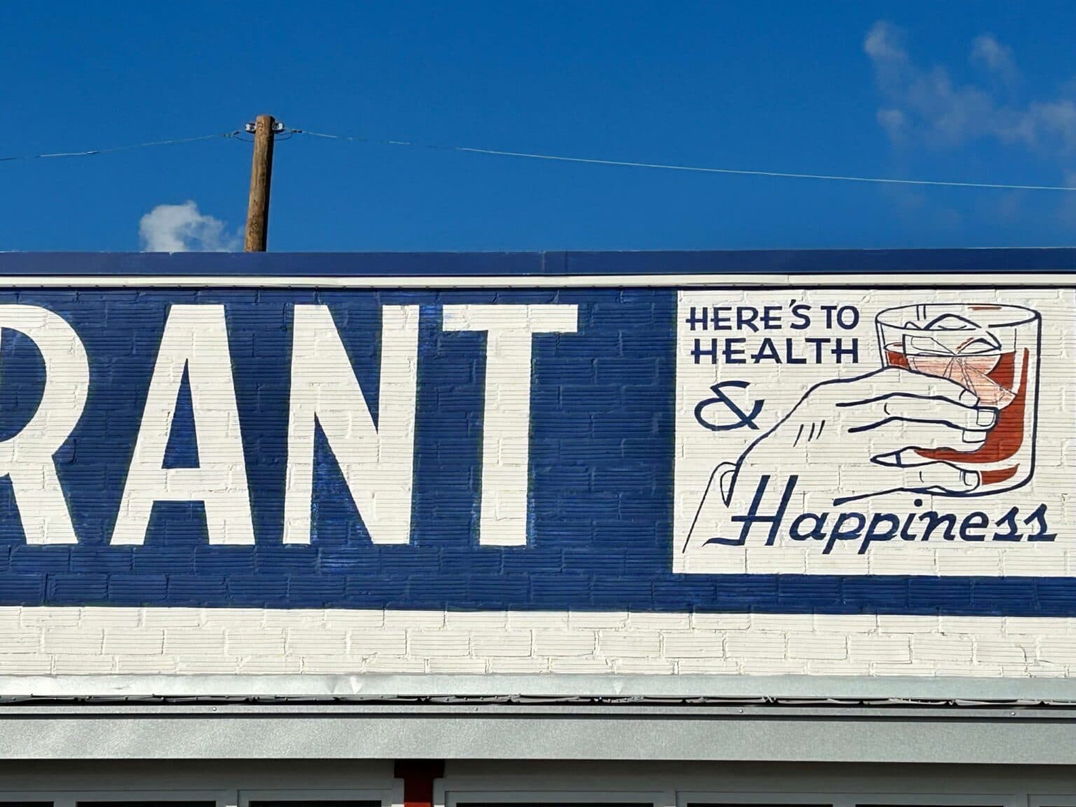 Sign on a restaurant featuring the phrase "Here's to Health & Happiness" alongside a hand holding a drink.