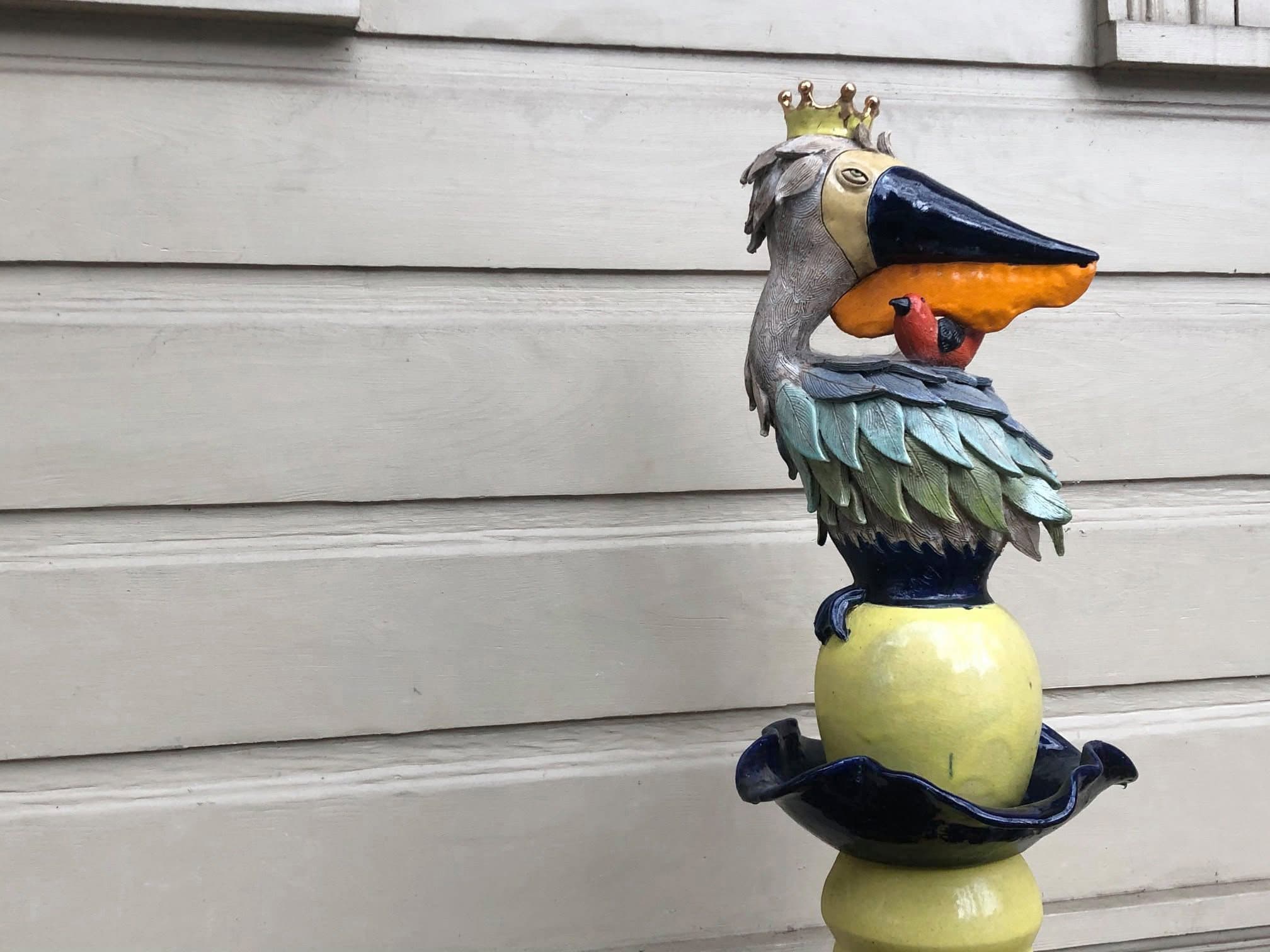 A whimsical ceramic sculpture of a crowned bird holding an orange fish, perched on a decorative base.