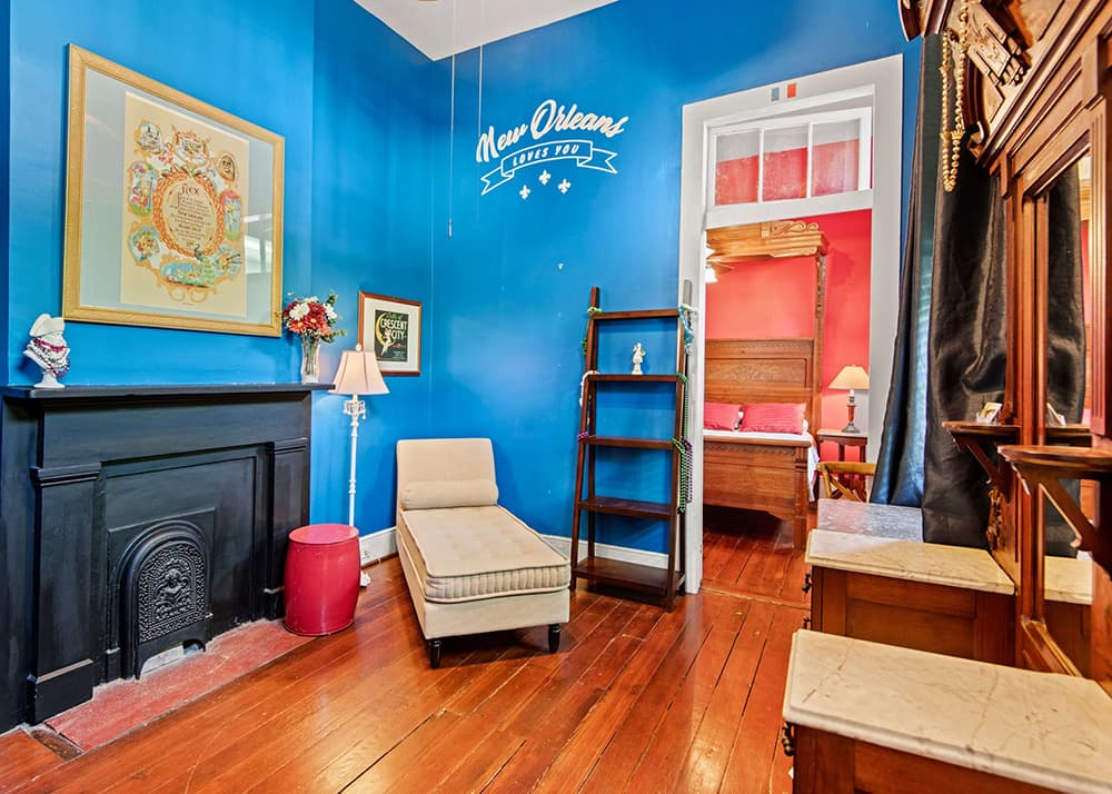 A cozy room with blue walls, a beige chaise lounge, a vintage fireplace, and a glimpse of a red bedroom through an open doorway.