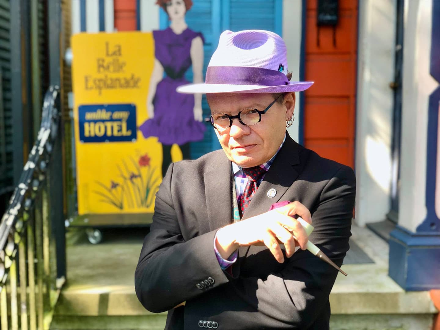 A person in a purple hat and suit stands confidently with crossed arms in front of a colorful building and a hotel sign.