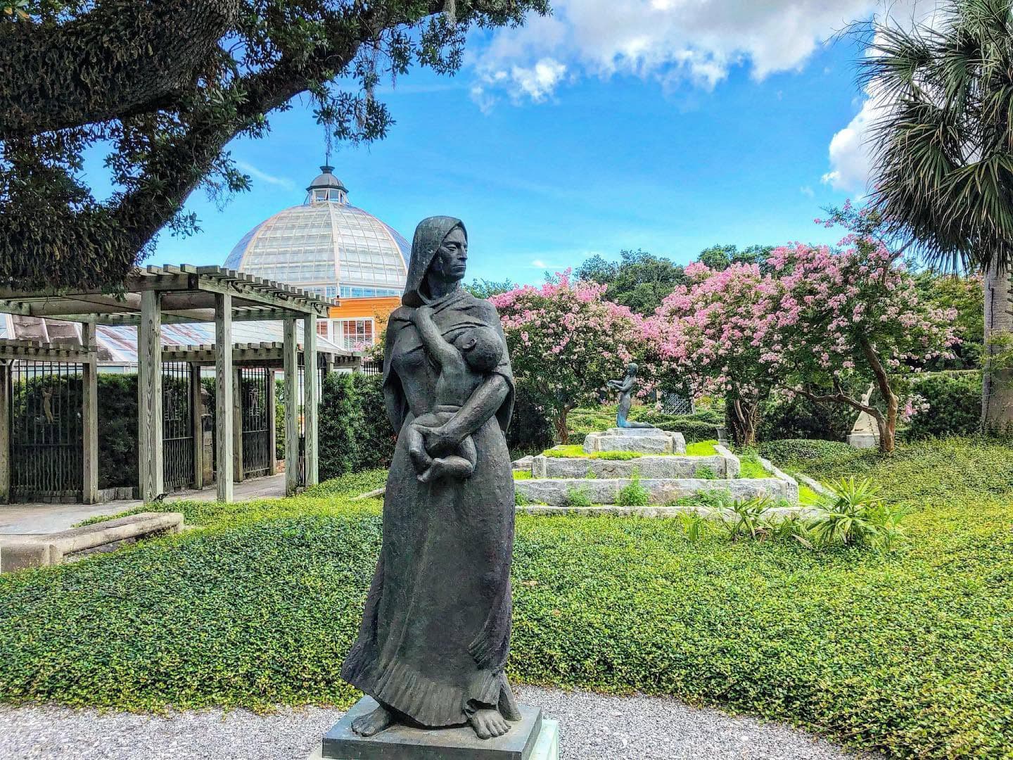 A bronze statue of a woman holding a child stands in a lush garden with flowering trees and a glass dome structure in the background.
