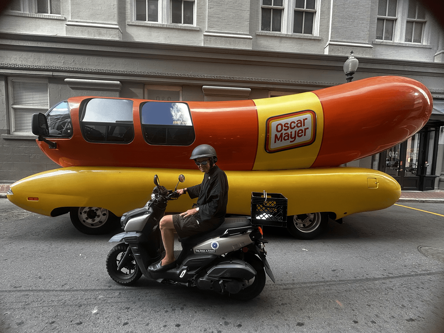 A scooter rider passes by the iconic Oscar Mayer Wienermobile parked on the street.