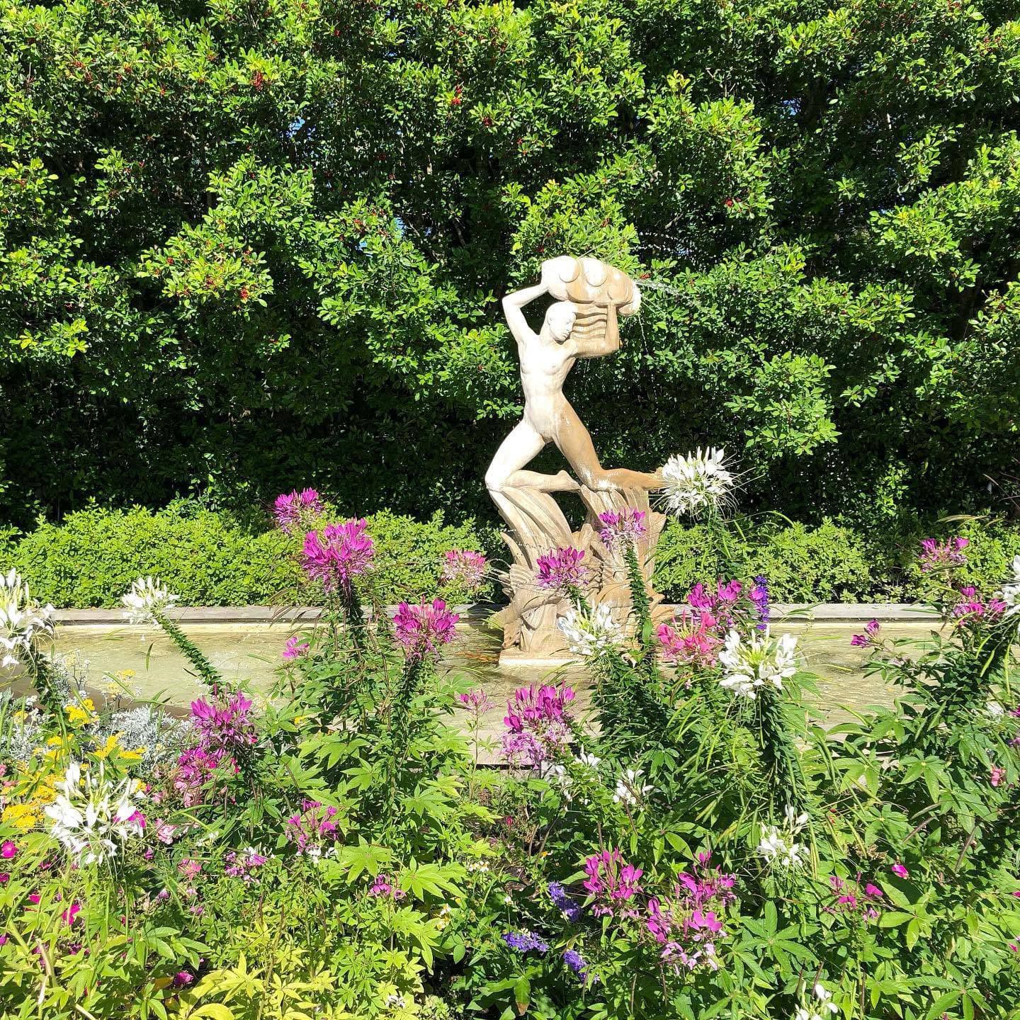 A nude sculpture of a woman holding a pot stands amidst vibrant flowers and greenery.