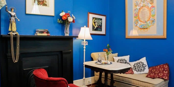 A cozy room with blue walls features a red armchair, a small table, a decorative fireplace, and colorful artwork.