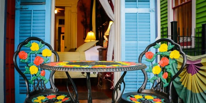 A colorful table and two chairs with floral designs sit outside an open blue door, revealing a cozy interior.