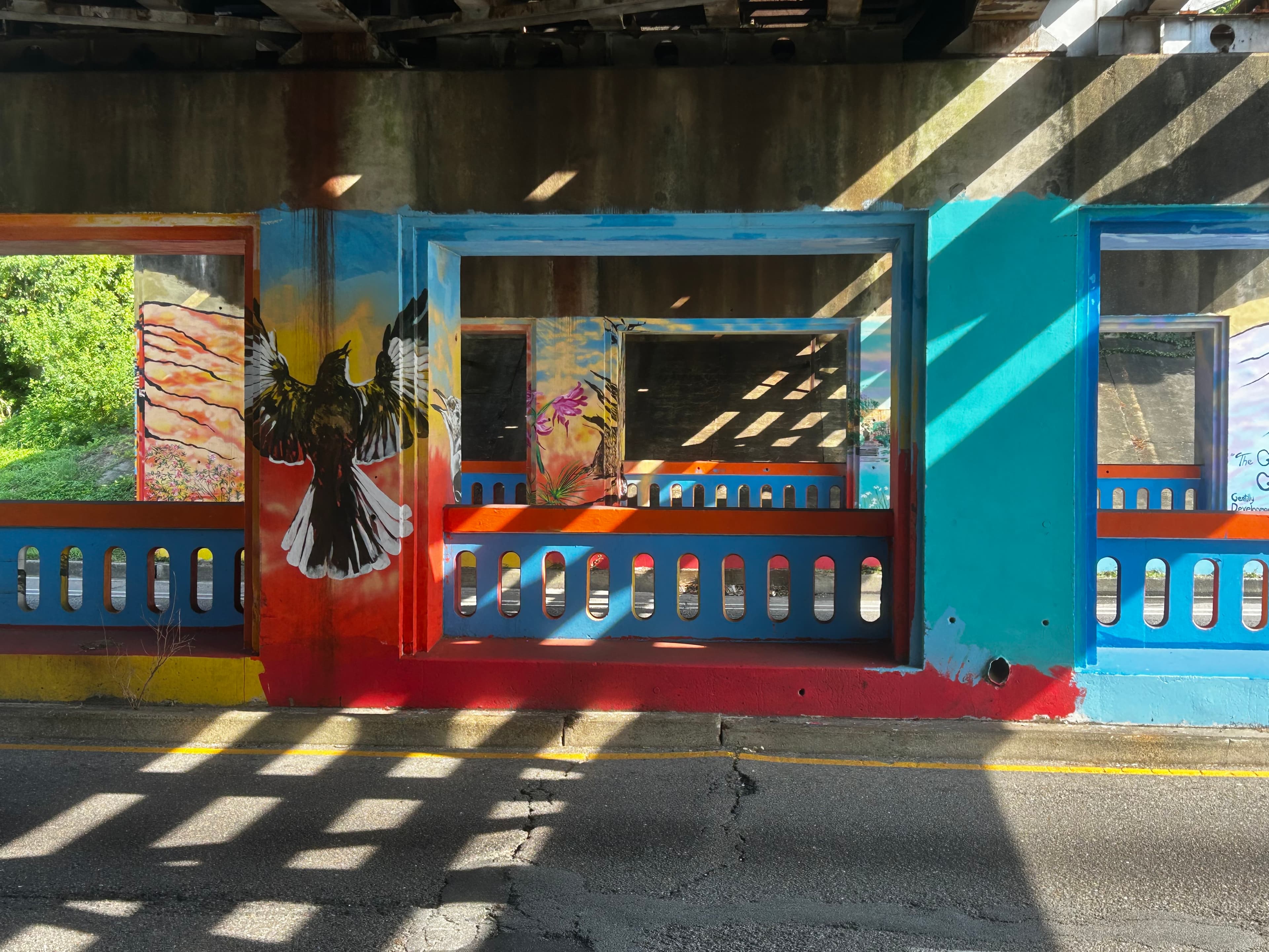 Colorful mural featuring a bird and abstract designs on an underpass wall, illuminated by sunlight.
