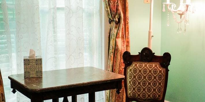 A small, elegantly furnished room featuring a decorative table and an ornate chair beside sheer curtains and a chandelier.