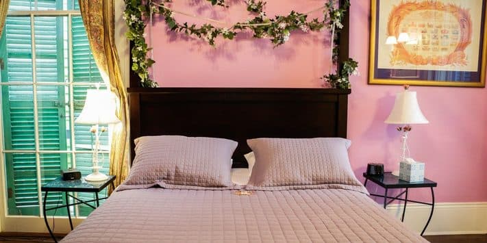 A cozy bedroom with a pink wall, a bed with lavender bedding, and lamps on either side.