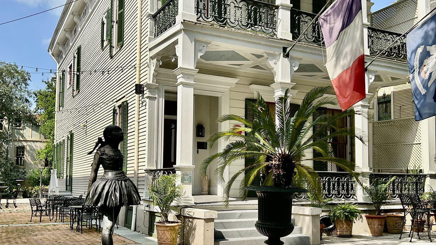 A bronze statue of a girl in a dress stands in front of a historic building with French and American flags.