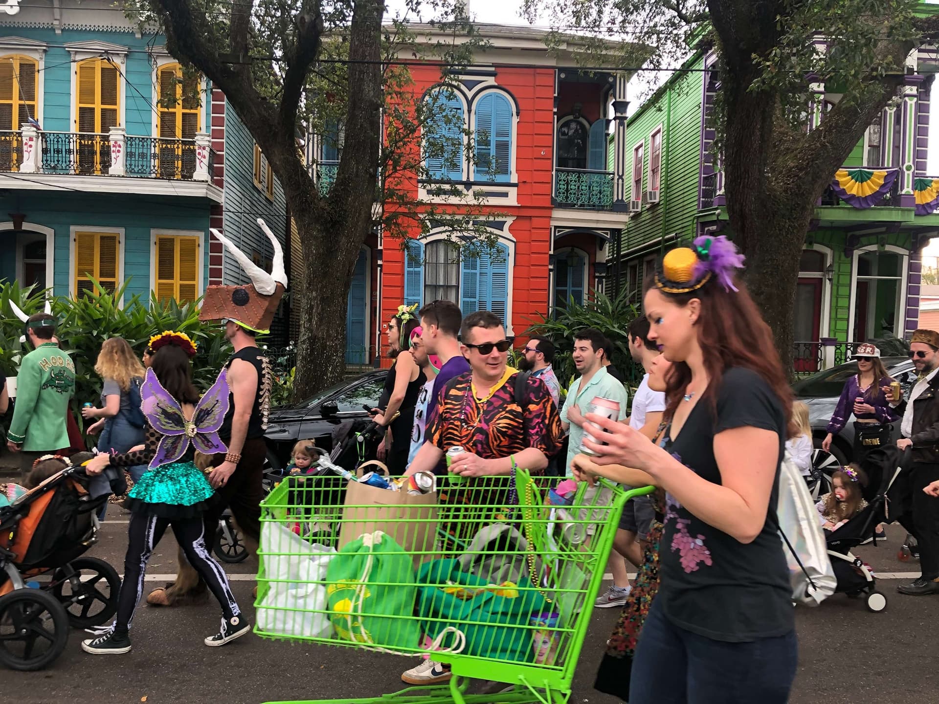 A lively street scene during a parade, featuring costumed individuals, strollers, and a green shopping cart in front of colorful houses.