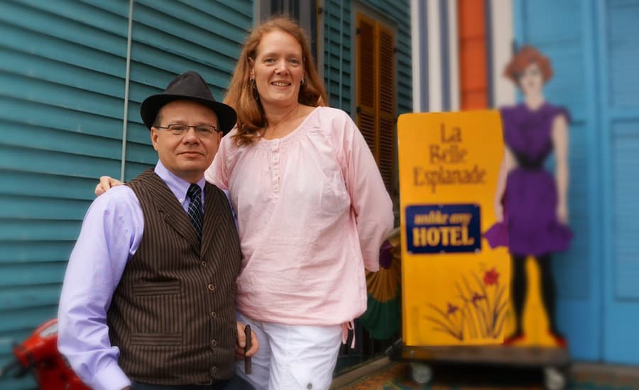 A man in a hat and a woman in a light blouse pose together in front of a colorful hotel sign.