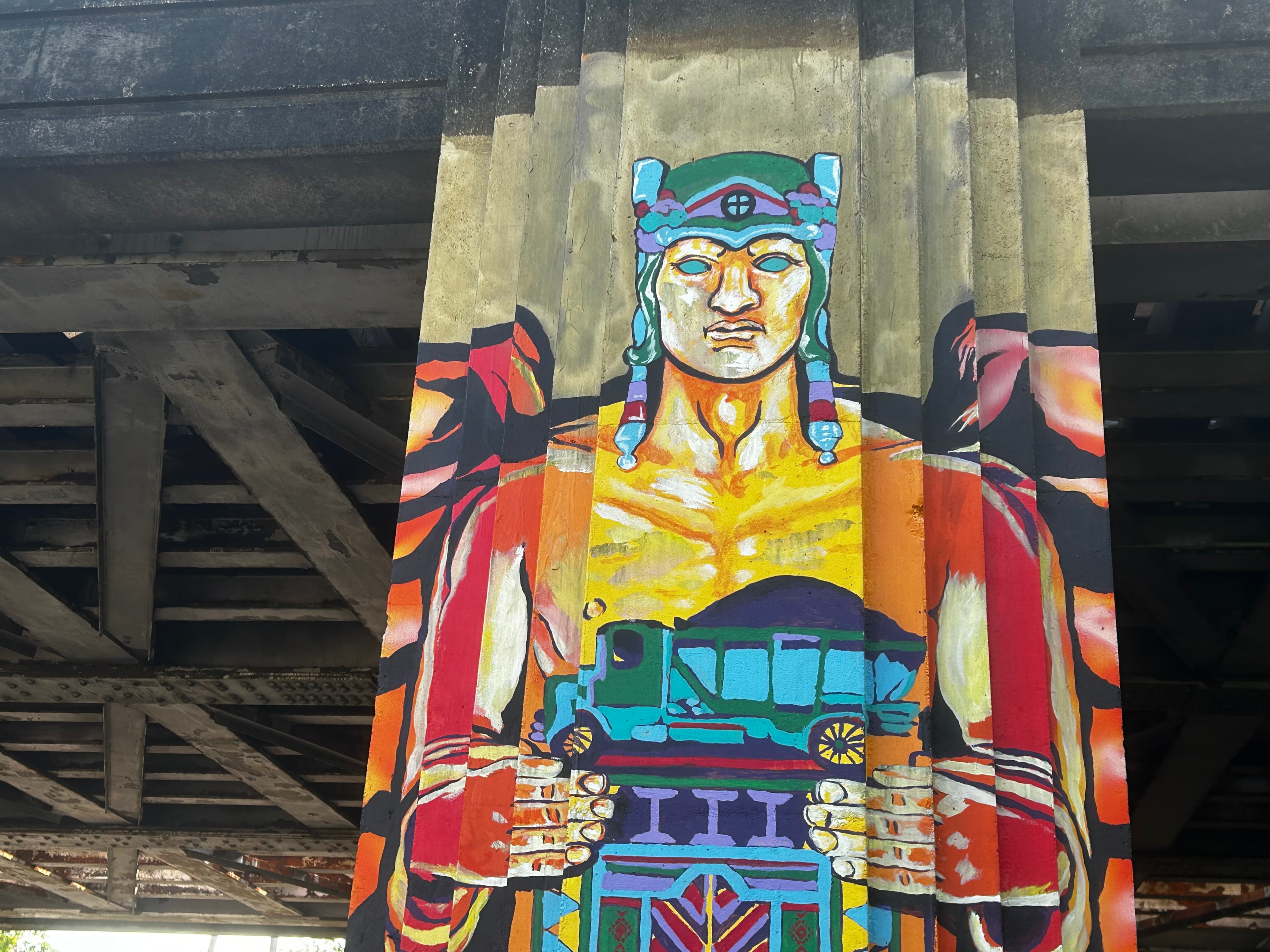 A vibrant mural of a muscular figure holding a small vehicle, situated on a bridge support. A vibrant mural of a muscular figure holding a small vehicle, situated on a bridge support.