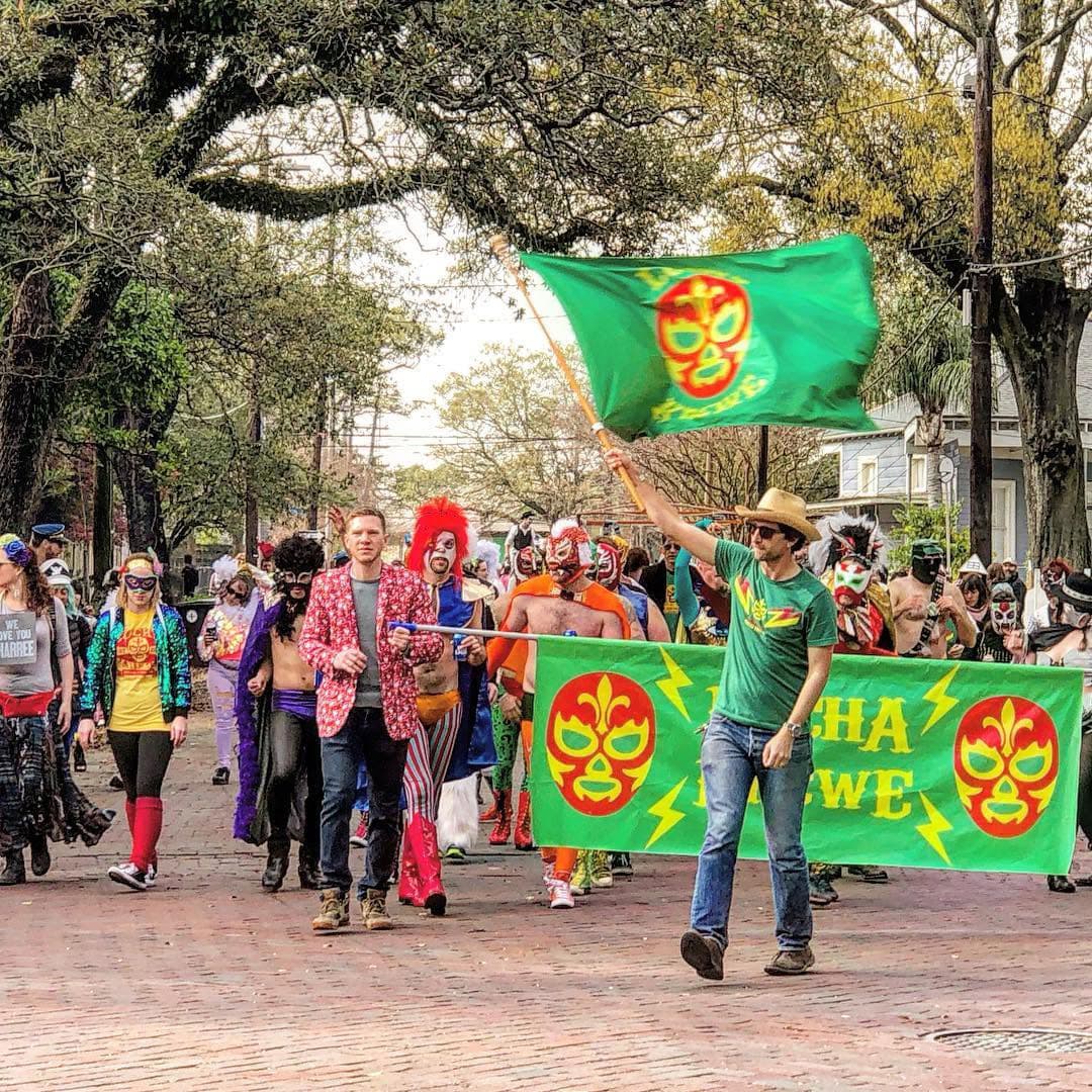 A lively parade featuring participants in colorful costumes, including clowns and luchador masks, holding flags and banners.