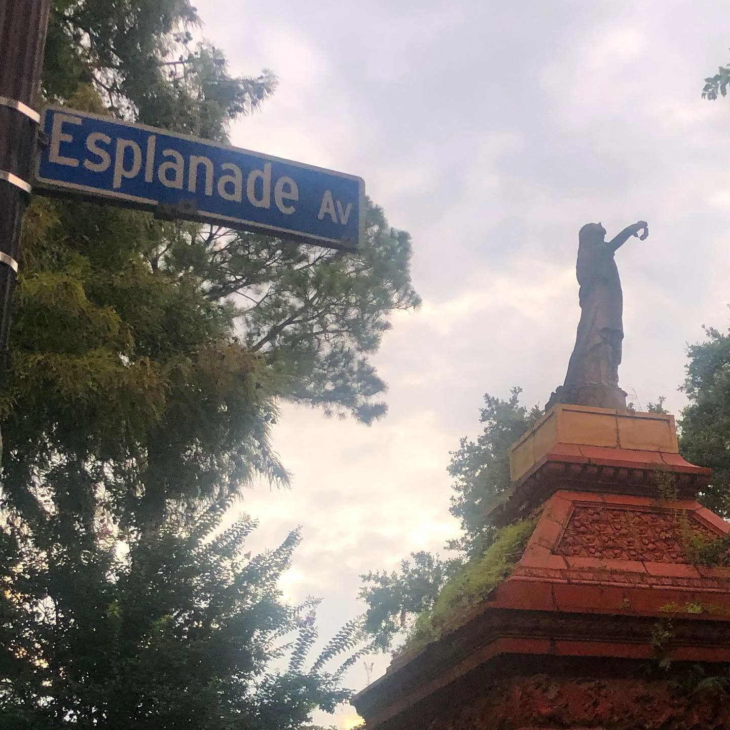 A statue stands atop a decorative pedestal beneath a street sign reading "Esplanade Ave" against a cloudy sky.
