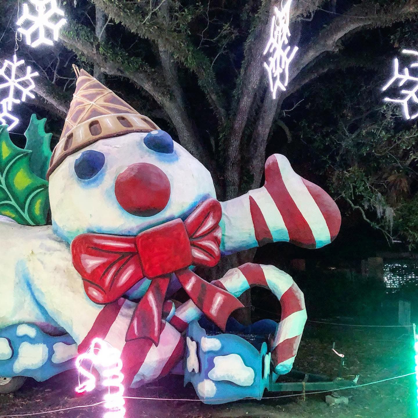 A colorful, whimsical snowman sculpture with an ice cream cone hat and a bow, surrounded by festive lights and decorations.