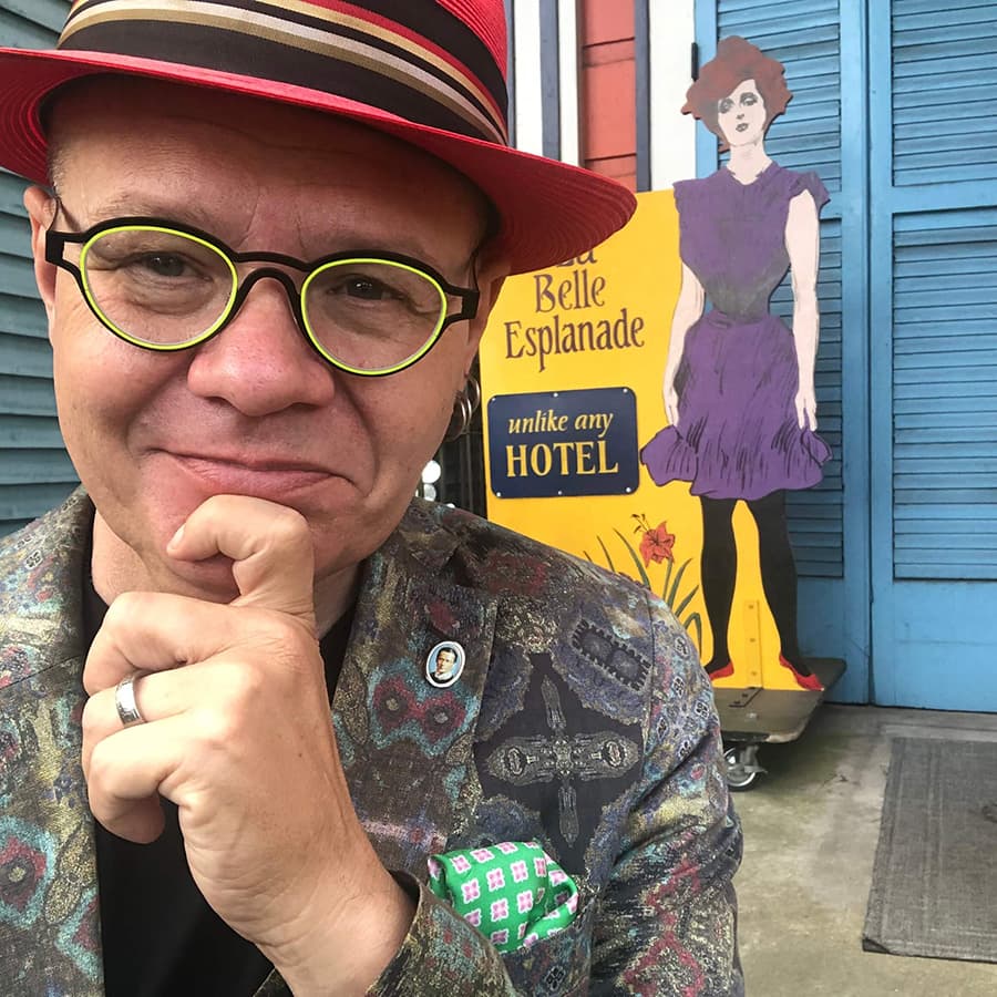 A person in a colorful hat and patterned jacket poses in front of a hotel sign featuring a woman in a purple dress.