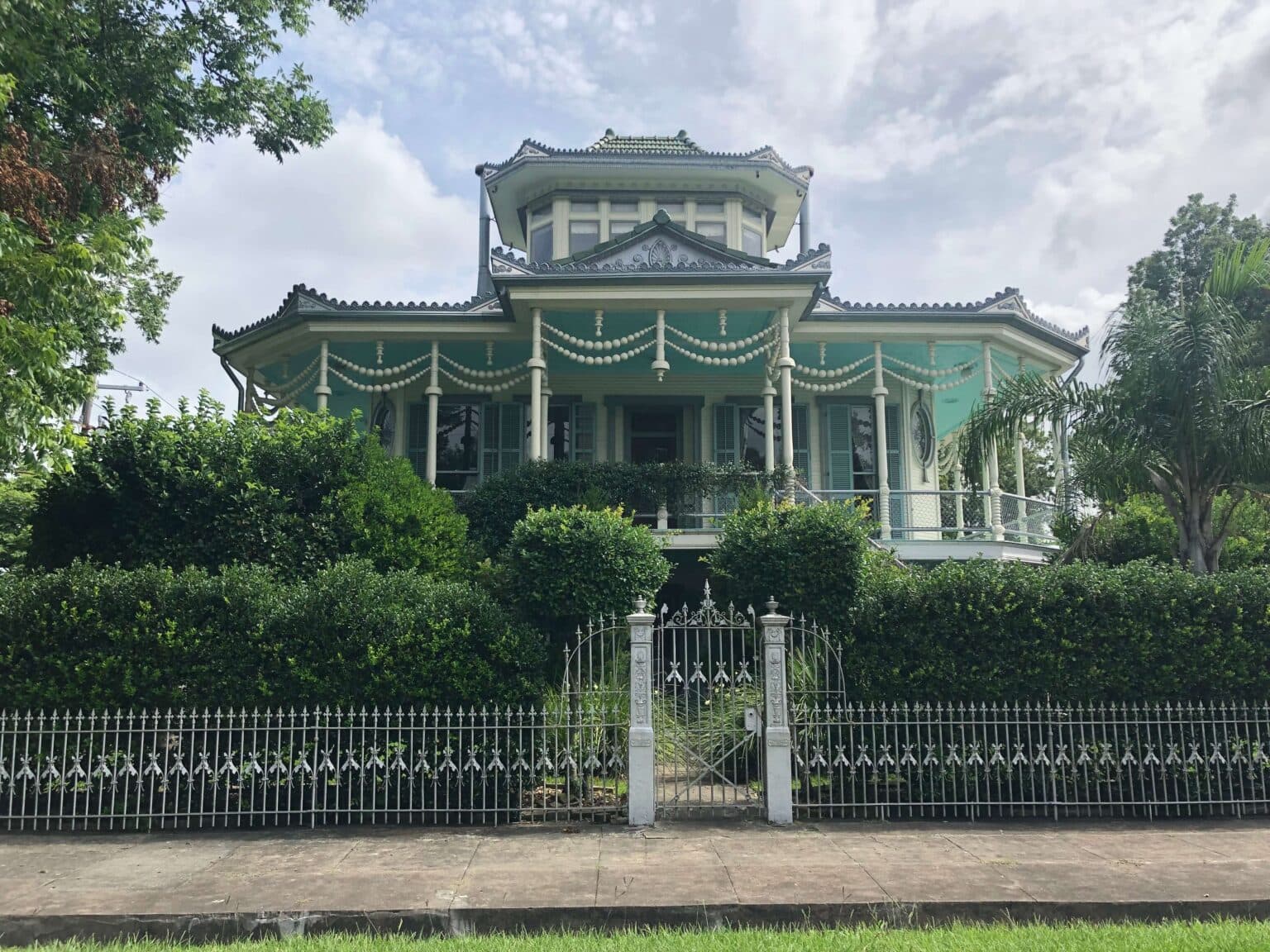 A vintage green house with decorative trim and lush gardens, enclosed by an ornate white fence.