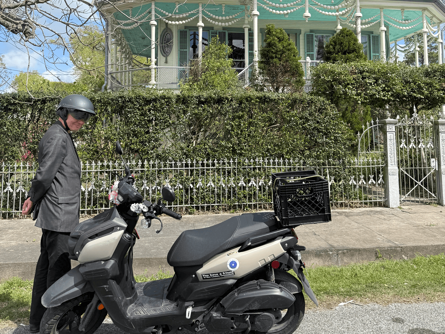 A person in a helmet stands beside a scooter in front of a decorative green house.