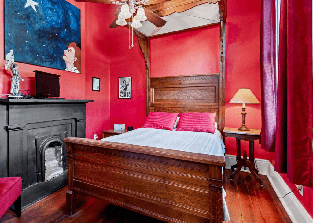 A cozy bedroom with a wooden bed, vibrant red walls, a lamp, and artwork, featuring a fireplace and a television.