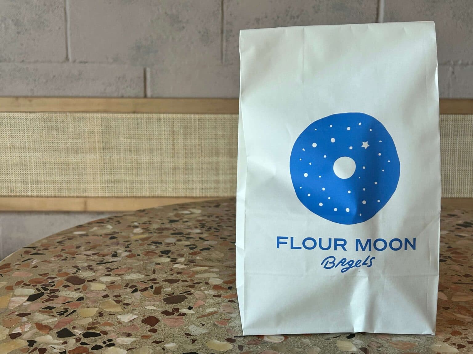 A white paper bag with a blue circular logo and text reading "FLOUR MOON Bagels" sits on a speckled table.