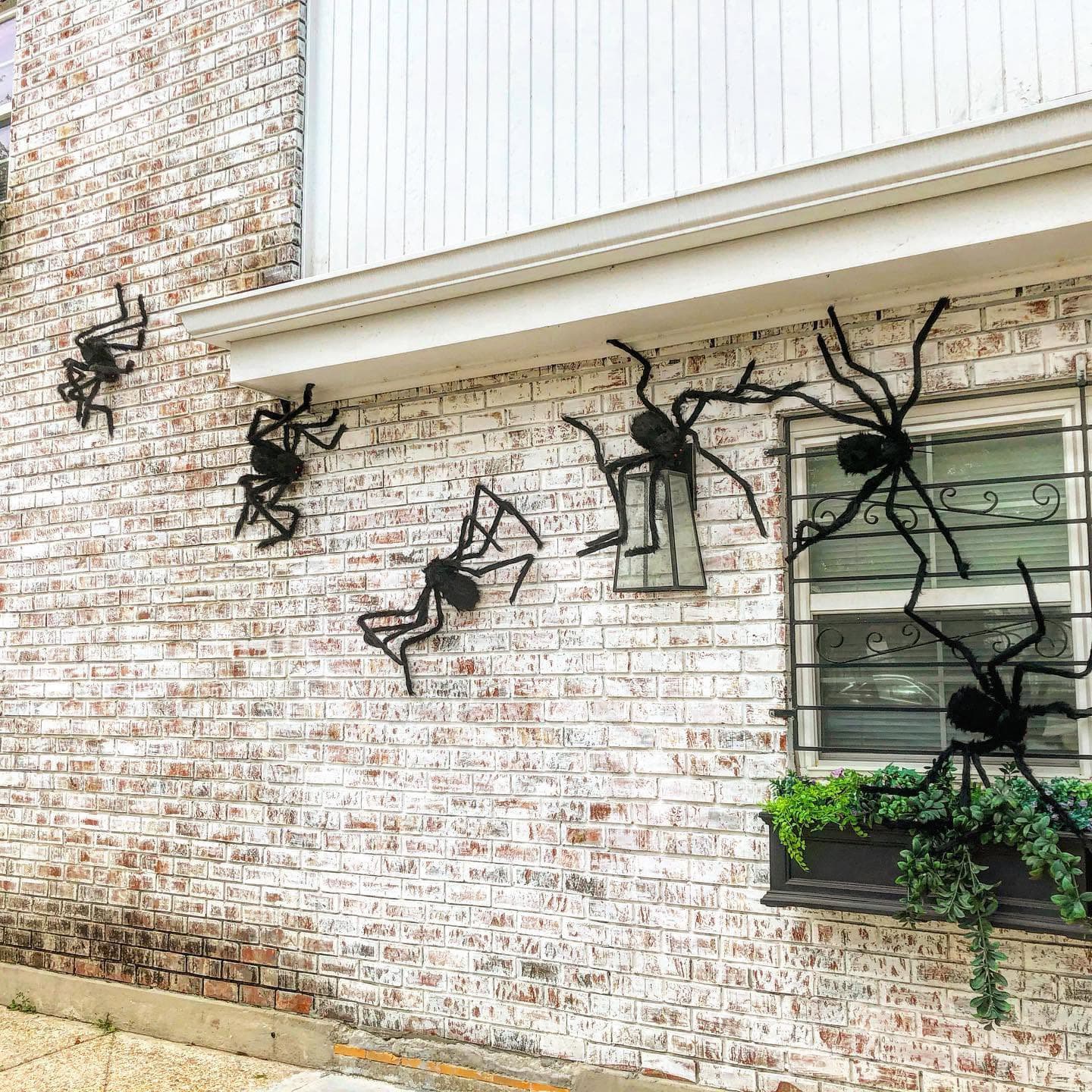 Large black spider decorations are attached to a brick wall near a window with greenery.