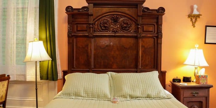 A cozy bedroom featuring a wooden bed with a decorative headboard and soft green bedding, accompanied by bedside lamps and curtains.