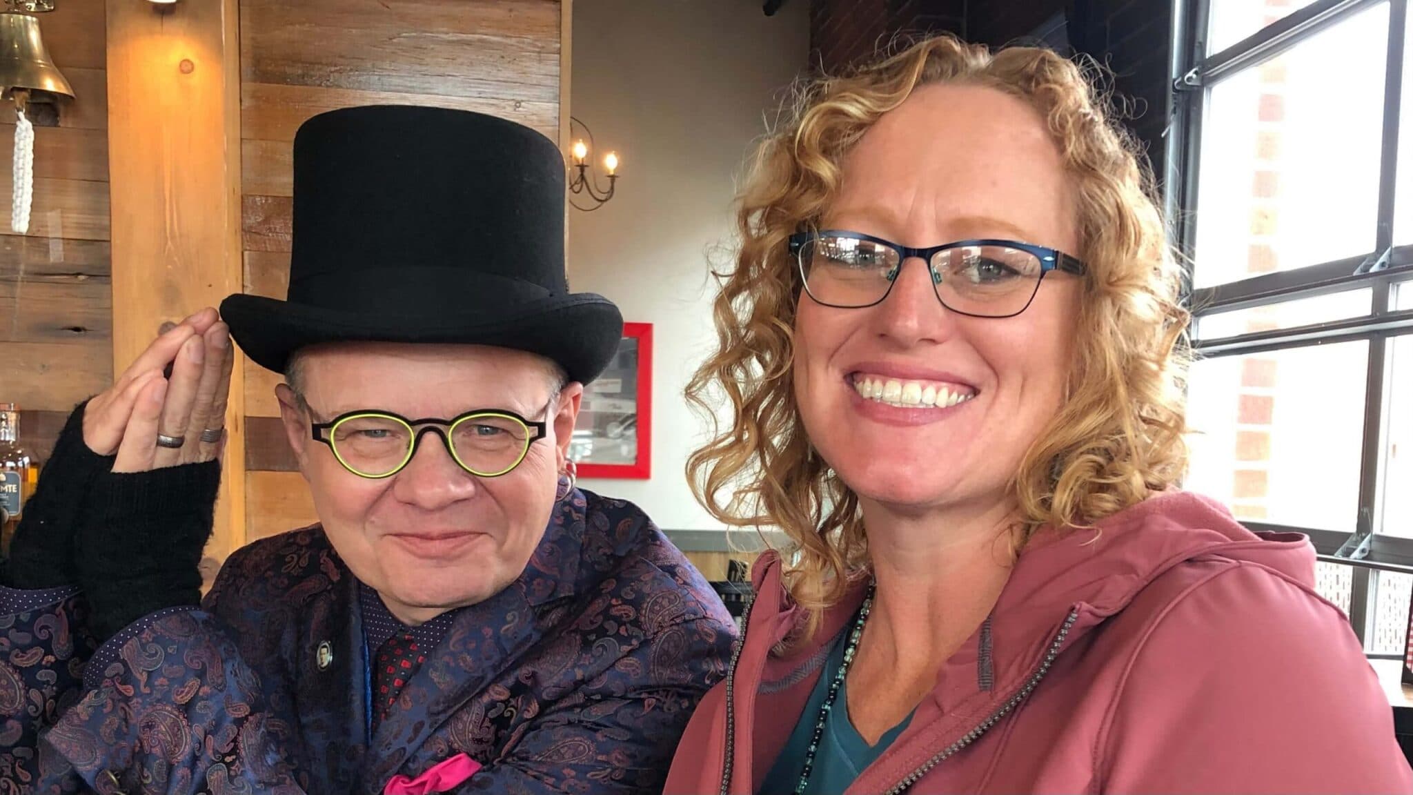 Two people are smiling together in a cozy restaurant setting, one wearing a top hat and colorful attire.