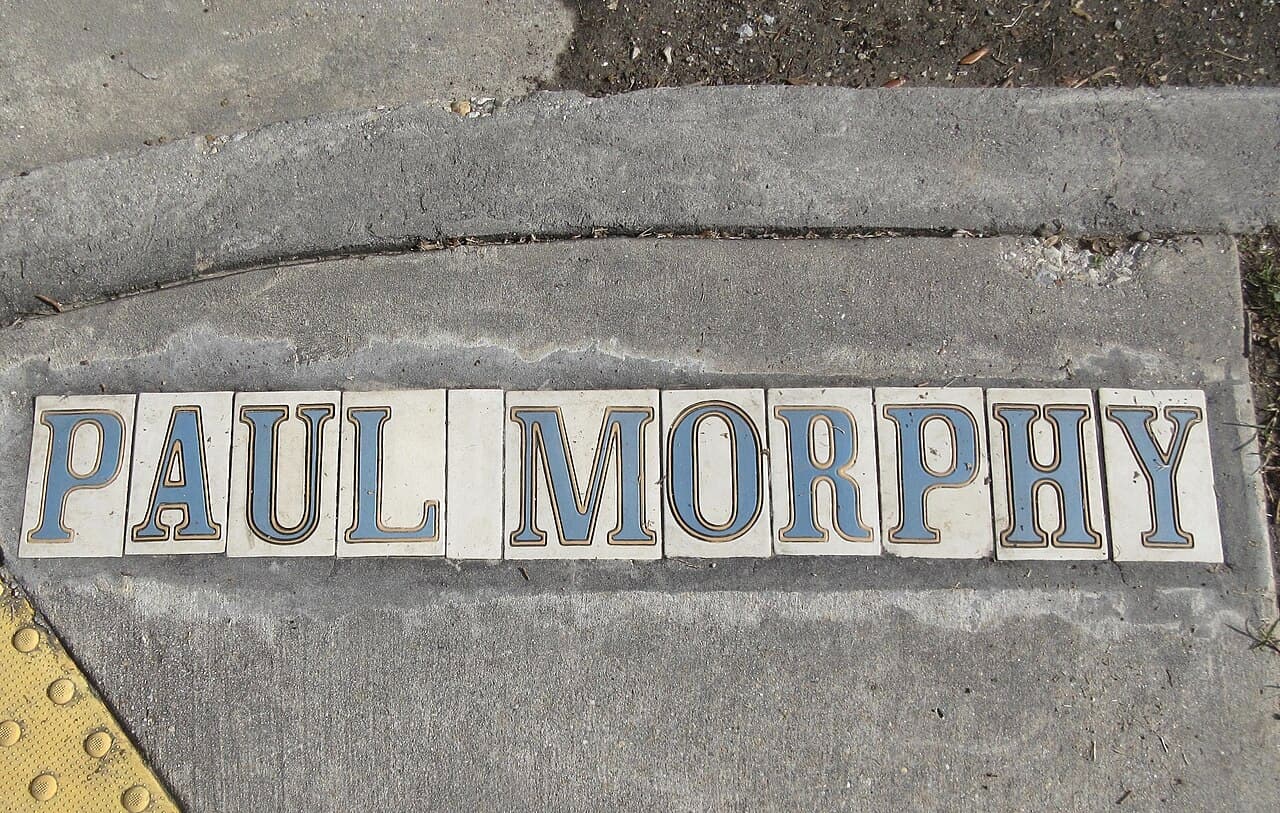 Sidewalk tiles spell out "PAUL MORPHY" in blue and gold letters on a concrete surface near a curb and a yellow textured section. Sidewalk tiles spell out "PAUL MORPHY" in blue and gold letters on a concrete surface near a curb and a yellow textured section.