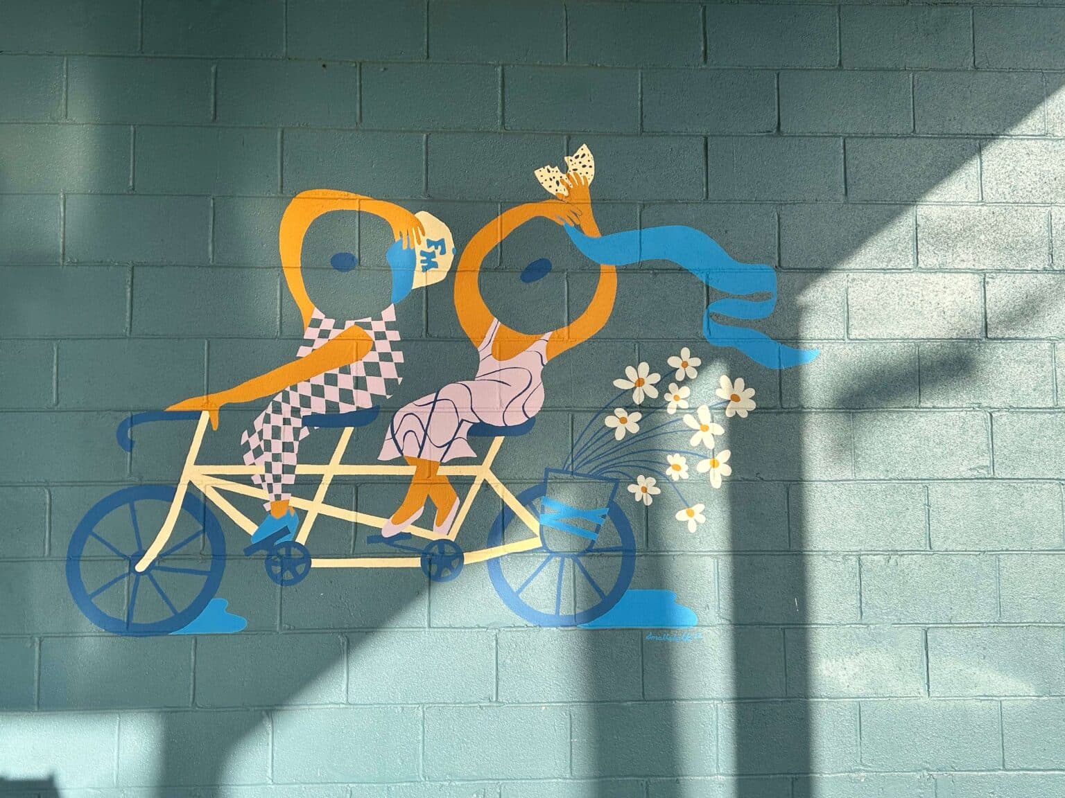A colorful mural depicting two playful figures riding a tandem bicycle with flowers and a flowing ribbon. A colorful mural depicting two playful figures riding a tandem bicycle with flowers and a flowing ribbon.