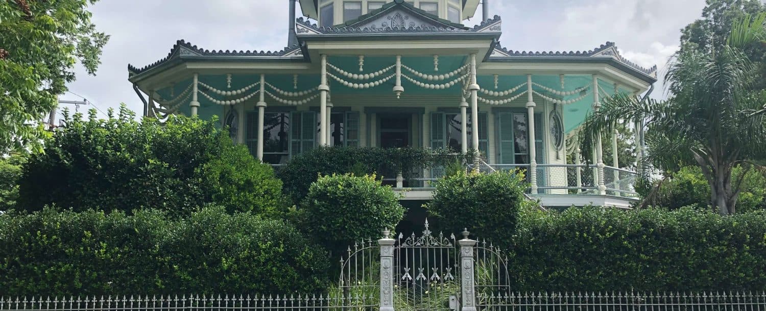 A Victorian-style house with green siding and decorative trim, surrounded by lush greenery and a white picket fence.